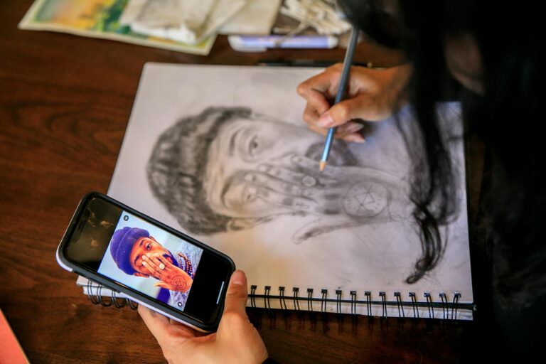 A person sketching a portrait using a smartphone reference on a spiral notebook.